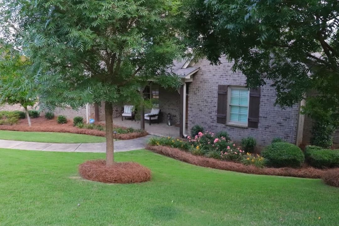 Brick house with shrubs and flowers, surrounded by a manicured lawn and trees.