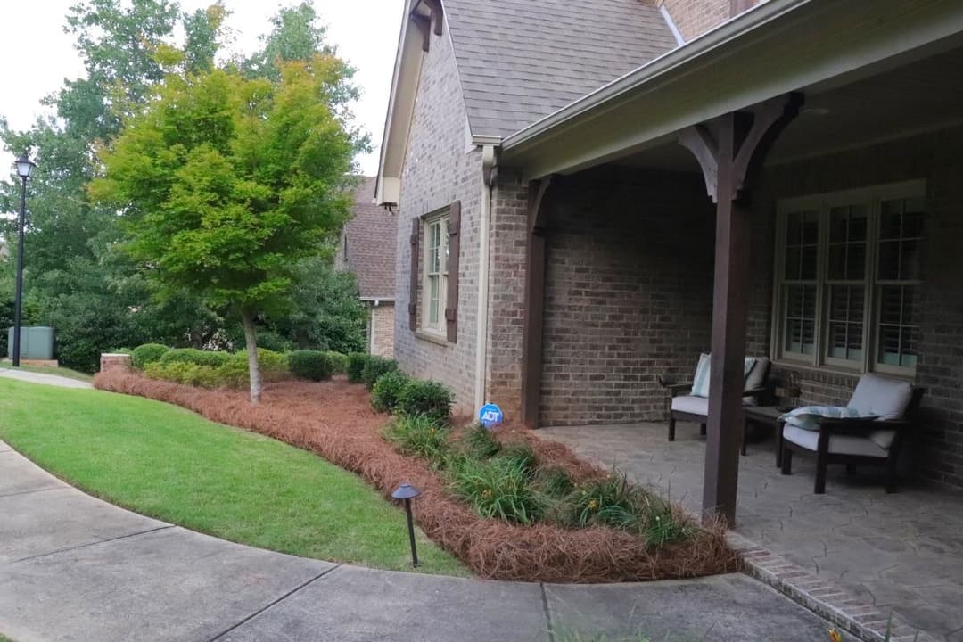 Front porch of a brick house with landscaped yard and seating area, surrounded by greenery.