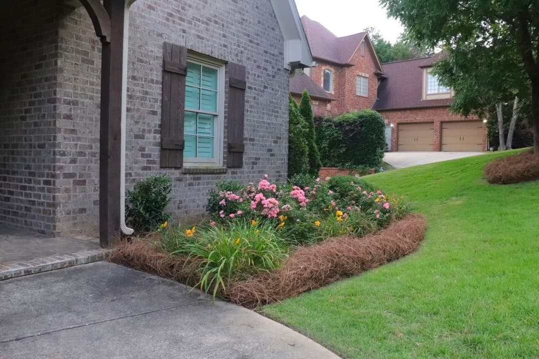 Lush garden with pink and yellow flowers beside a brick house entrance and driveway.